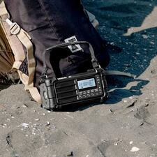 zwart Digitale survivalradio staat in het zand naast een rugzak met een drager op de rug.