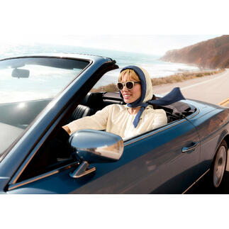 Vrouw in een cabrio met hoofddoek en zonnebril rijdt langs de kust.