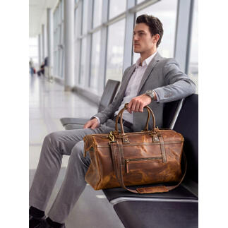 Man zit op de luchthaven met een buffellederen weekendtas naast hem.