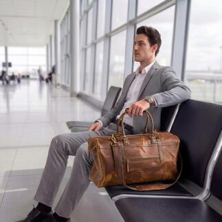 Man zit op de luchthaven met een buffellederen weekendtas naast hem.