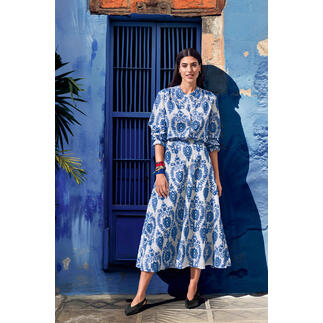 Vrouw in een blauwe jurk met wit patroon, staand voor een blauwe muur.