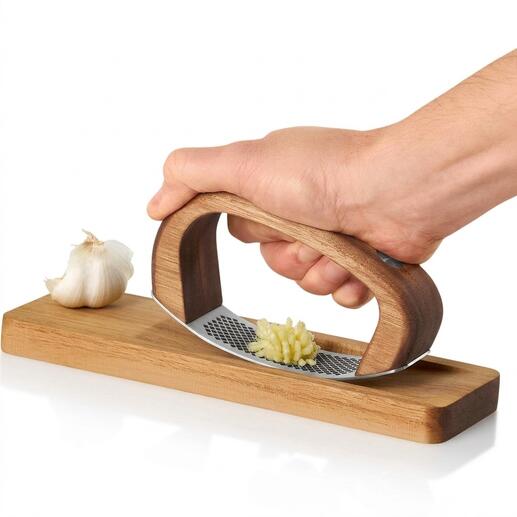 Hand houdt AdHoc knoflookpers boven houten plank met een teentje knoflook.