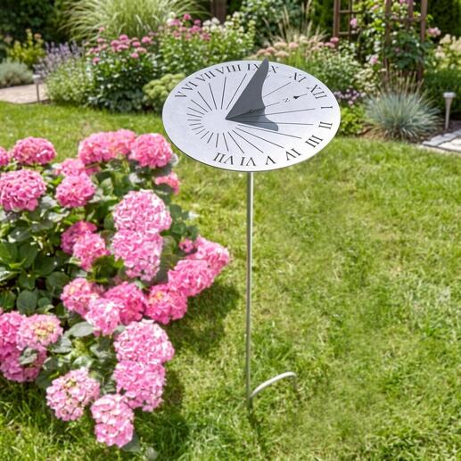 RVS zonnewijzer op een groen gazon, omgeven door roze bloemen.