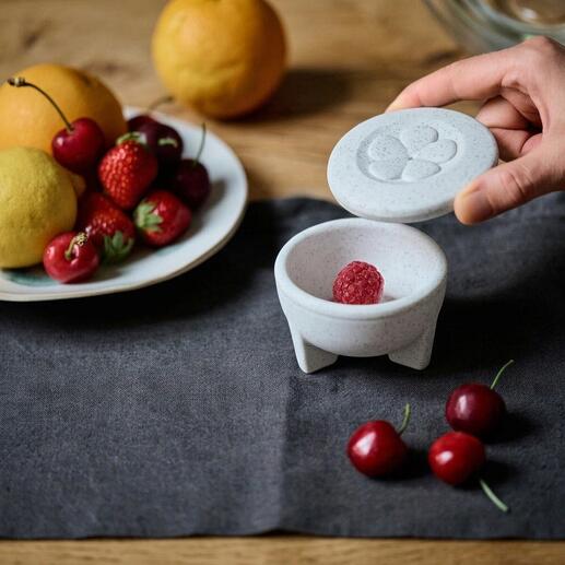 Porseleinen fruitvliegenval met een hand die het deksel opent en fruit op tafel.