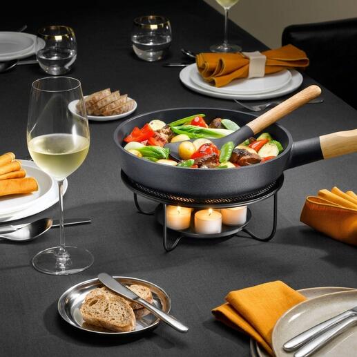 Pan met groenten op een vuurplaat, naast een wijnglas op een gedekte tafel.