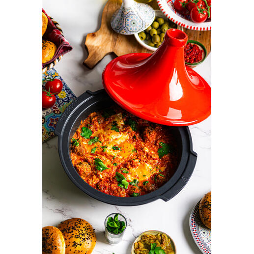 Tajine Peugeot Saveurs met een rode deksel, gevuld met kleurrijke groenten en kruiden, op een tafel met bijgerechten.