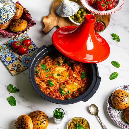 Tajine Peugeot Saveurs met een rode deksel, gevuld met kleurrijke groenten en kruiden, op een tafel met bijgerechten.