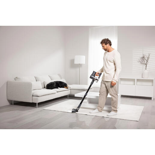 Man zuigt met een draadloze stofzuiger op een tapijt, een hond ligt op een bank.