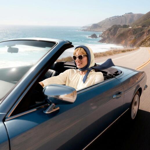 Vrouw in een cabrio met hoofddoek en zonnebril rijdt langs de kust.