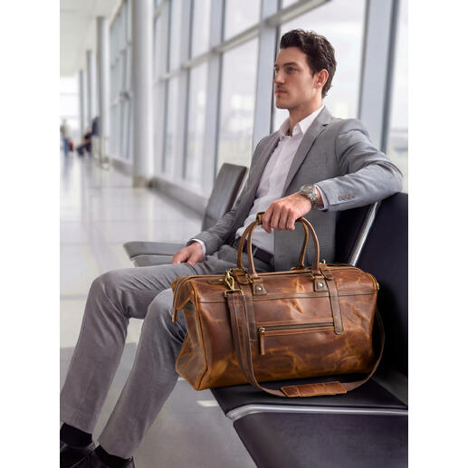 Man zit op de luchthaven met een buffellederen weekendtas naast hem.