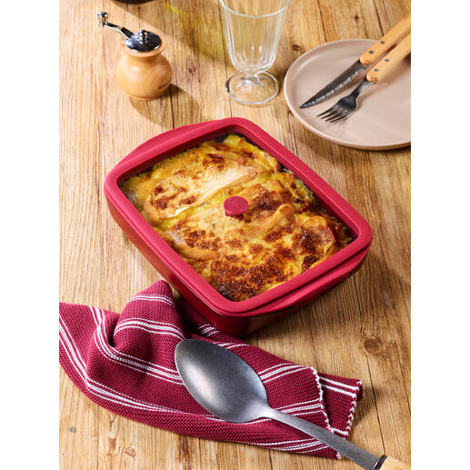 Rode ovenschaal met deksel, gevuld met bonenschotel, op een houten tafel.