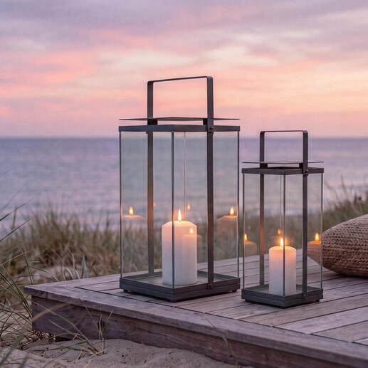 Twee design-laternen op een houten terras aan het strand bij zonsondergang.