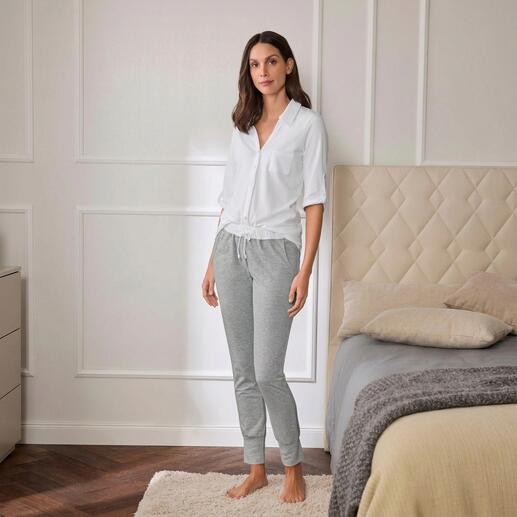 Vrouw in een witte pyjama-top en grijze joggingschoenen staat naast een bed.