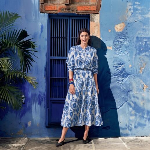Vrouw in een blauwe jurk met wit patroon, staand voor een blauwe muur.