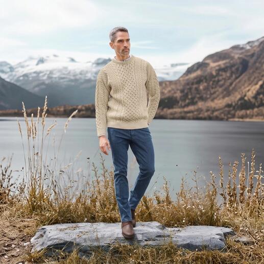 Man draagt een Merino-Aran-trui in wolwit, staande voor een meer met besneeuwde bergen op de achtergrond.
