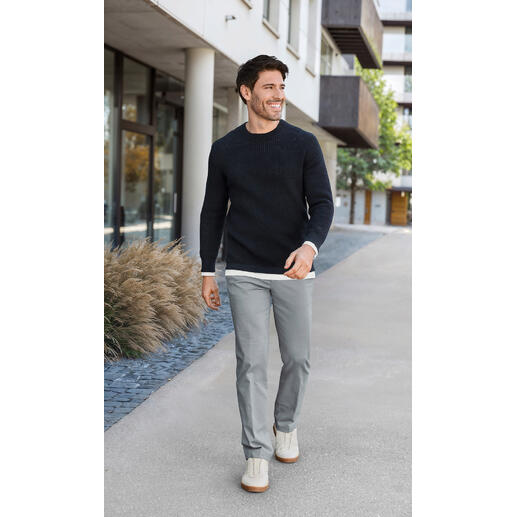 Hiltl oxford-chino Man in een zwarte wafeltrui en grijze broek, loopt over een moderne stoep.