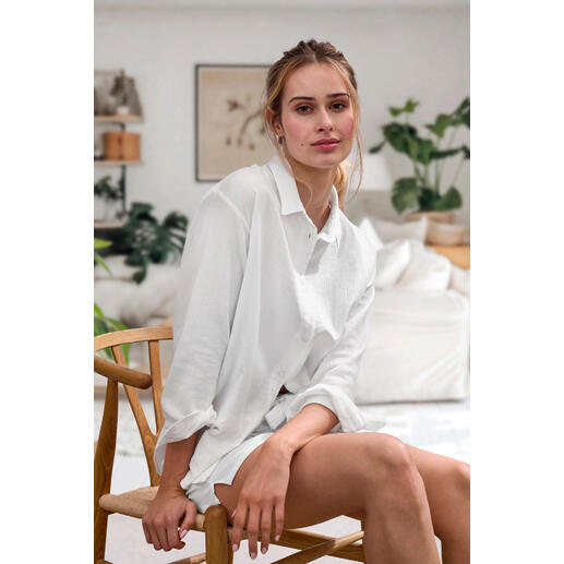 blouse en short Vrouw zit op een stoel, draagt een witte Novila Musselin blouse, met natuurlijke planten op de achtergrond.