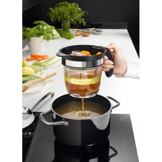 Hand met een zeef giet groentebouillon in een pan, terwijl een keuken op de achtergrond zichtbaar is.