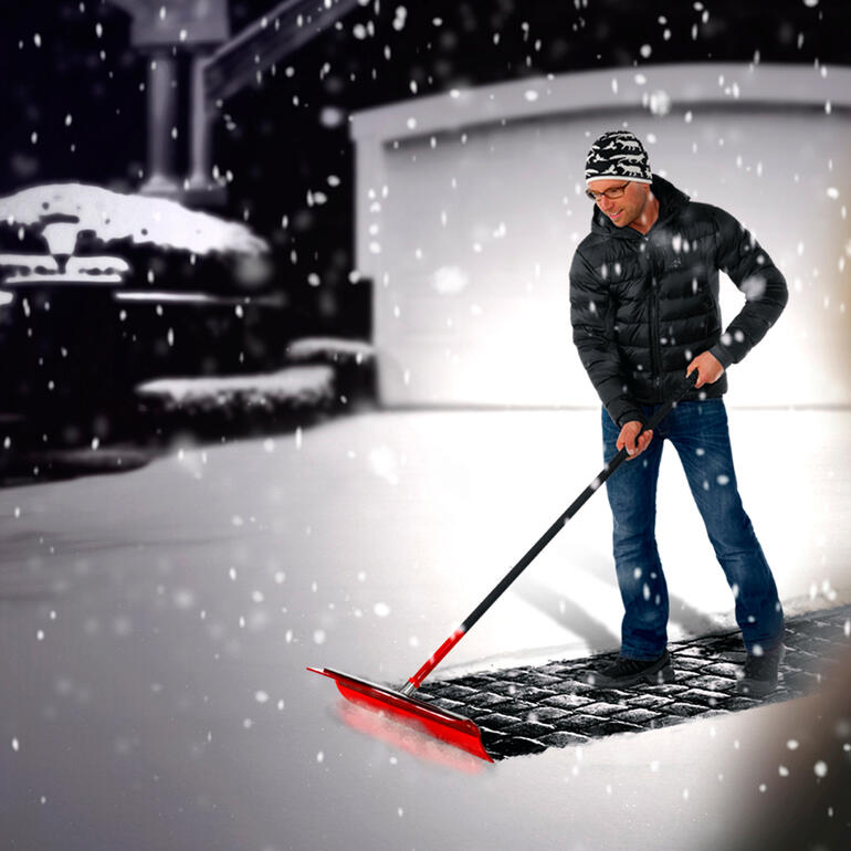 Man schept sneeuw met Schneeade® voor een garage in een besneeuwde omgeving.
