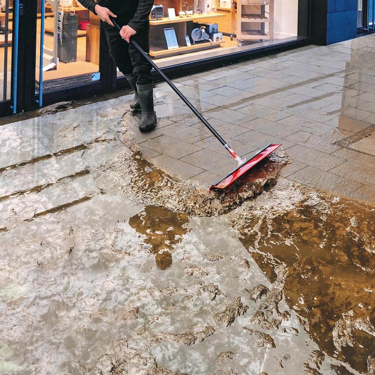 Man met bezem die water van het trottoir veegt.