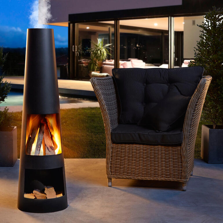 Moderne terrasbrander met brandend hout naast een comfortabele stoel.