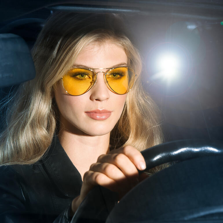Vrouw met gele nachtbrilclip in de bestuurdersstoel van een auto.