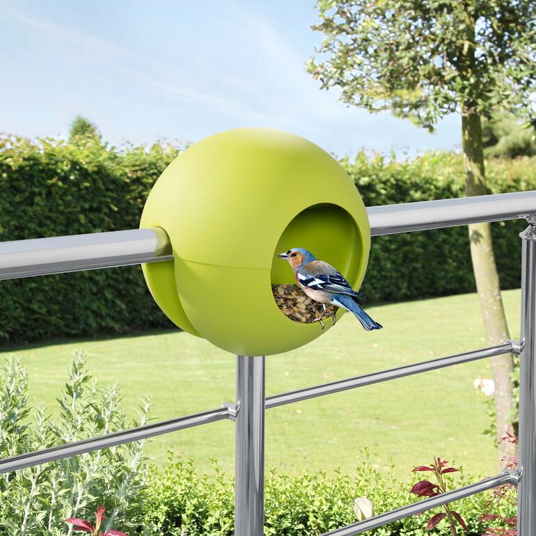 Vogelhuisje van groen materiaal, bevestigd aan een balkonleuning met een vogel die voert.
