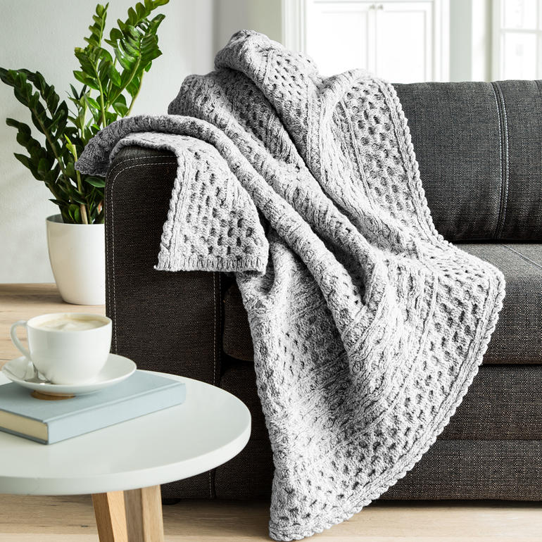 Aran gebreide plaid in lichtgrijs, over een bank gedrapeerd, met een kopje koffie.