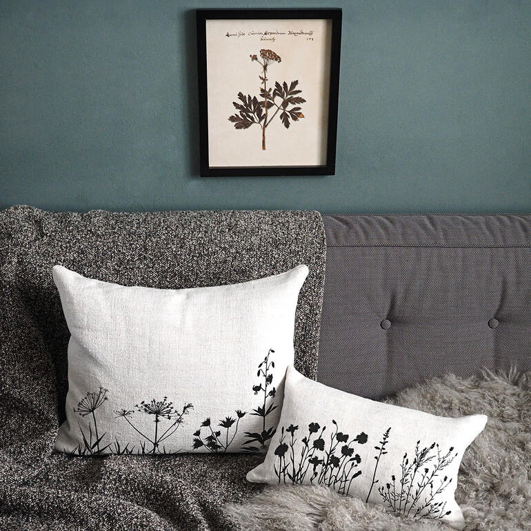 Het dessin van botanische motieven is afkomstig van een atelier uit Hannover, waar alles nog strikt met de hand wordt verwerkt. Twee linnen kussens met zwarte plantenmotieven op een bank, een schilderij hangt erboven.