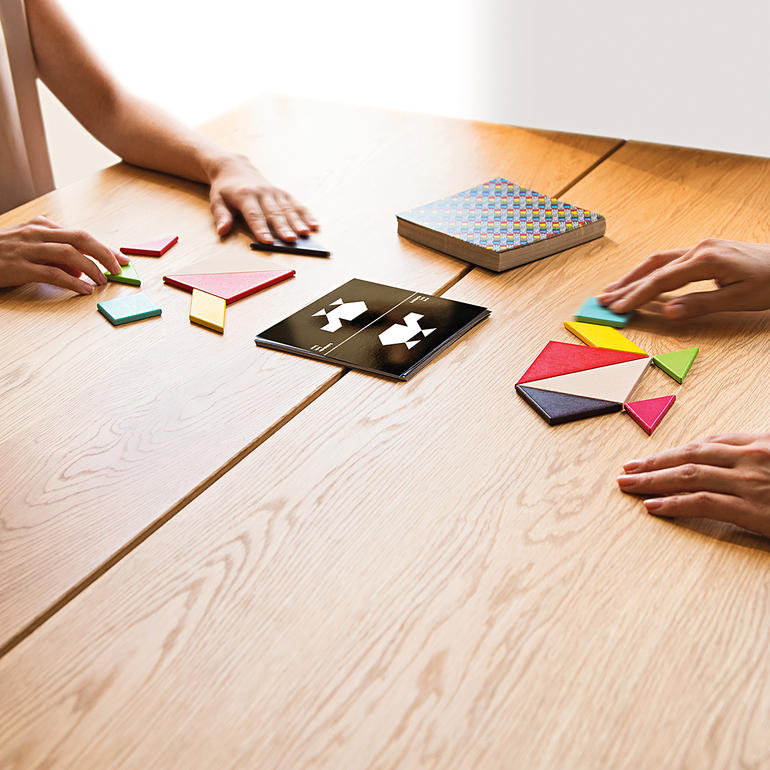 Twee personen spelen Tangram aan een tafel met houten oppervlak.