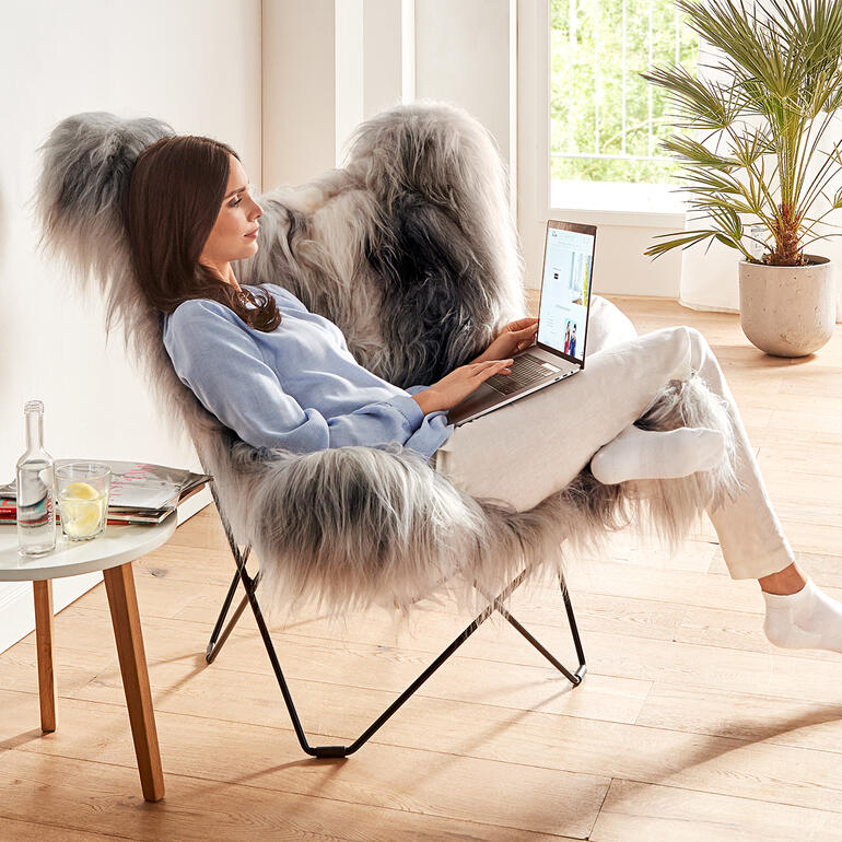 De rugleuning van deze fauteuil heeft een royale breedte en hoogte, en zit heerlijk comfortabel, ook voor grotere personen. Vrouw zit in de Mariposa loungestoel met laptop, lichte kamer met planten.