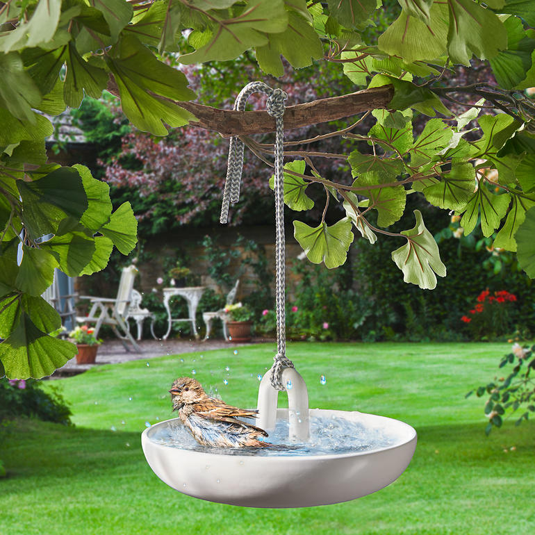 Hangend vogelbad van wit materiaal, opgehangen aan een tak.