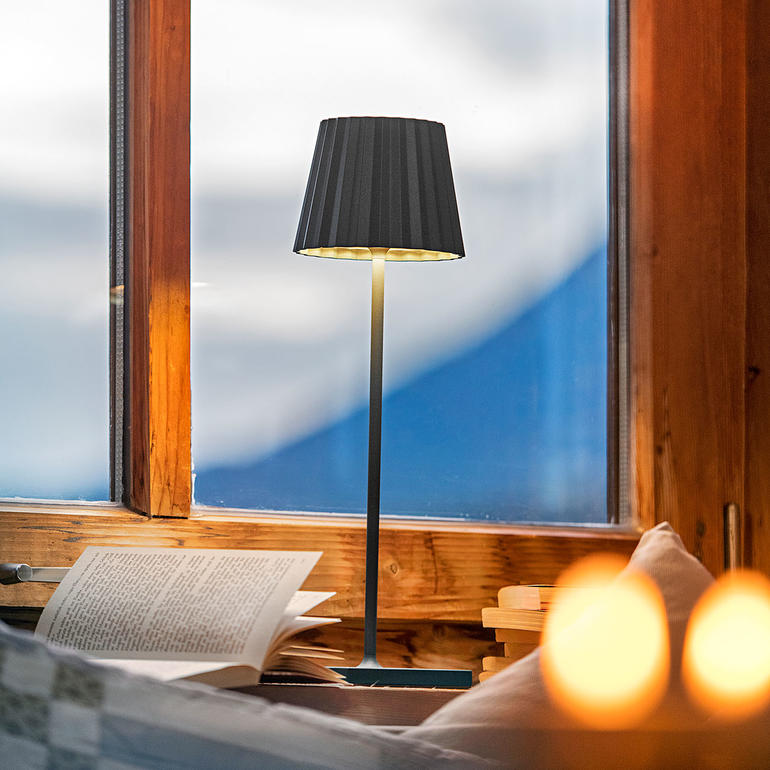 Oplaadbare tafellamp in zwart, op een tafel naast een vensterbank.