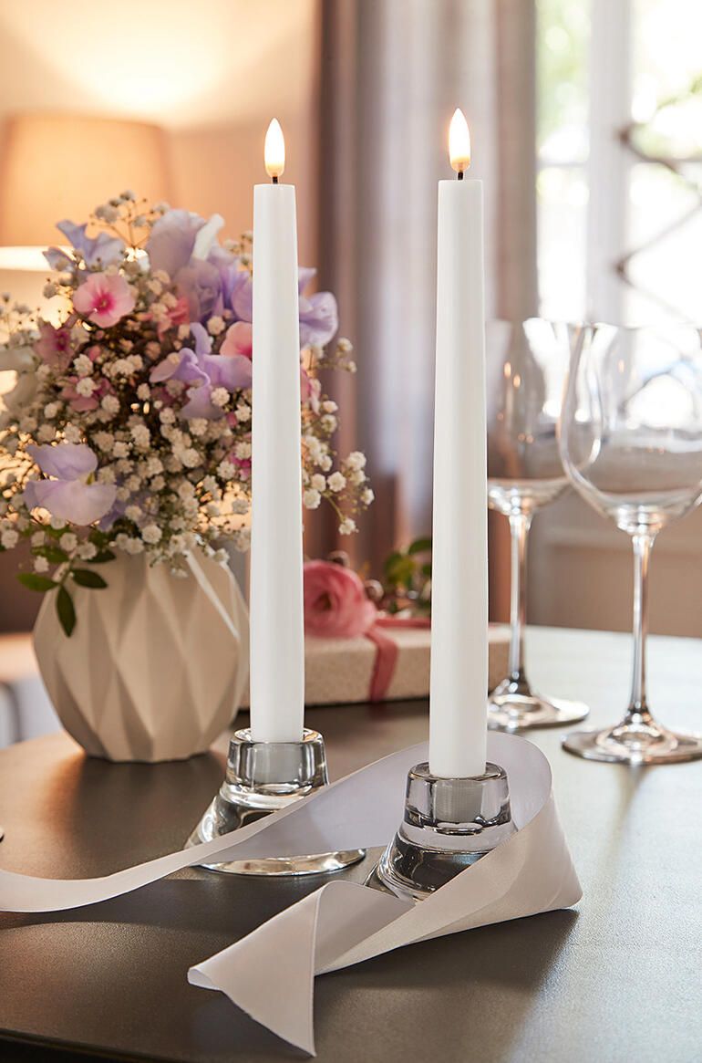 Twee LED-stabkaarsen in een decoratieve omgeving naast bloemen.