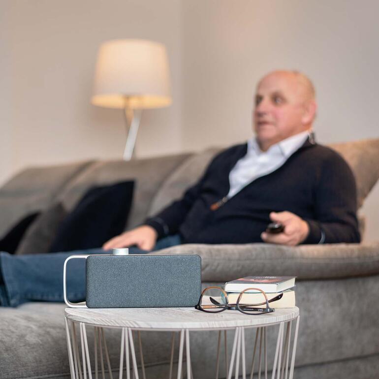 Oude man zit op een bank, kijkt televisie en gebruikt de TV-hoorversterker OSKAR.