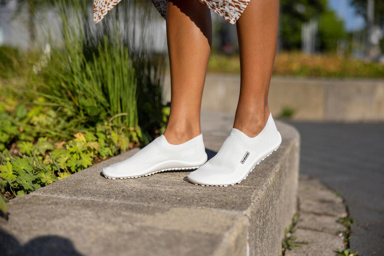 Witte Barfuß-leguano® Slipper op de vloer, met een vrouw op de achtergrond.