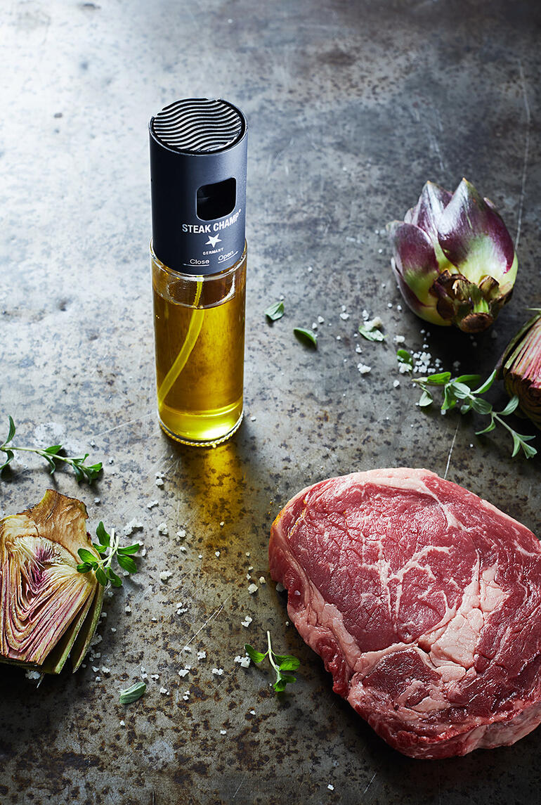 Olie-spuitfles met filter naast rauwe steak en kunstbloemen op een rustieke tafel.