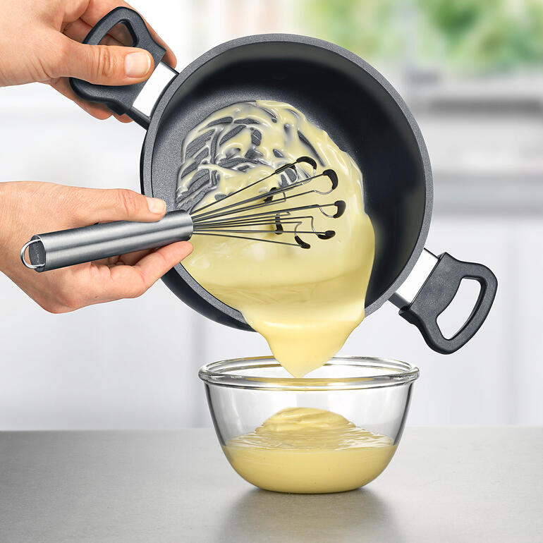 U bereikt alle ingrediënten zonder restjes achter te laten – helemaal tot aan de rand van de pan. Hand die deeg uit een pan in een kom giet