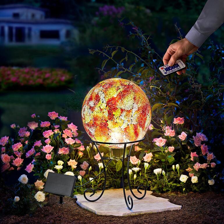 Zonne-energie tuinbal op een standaard, omringd door bloemen, hand houdt afstandsbediening vast.