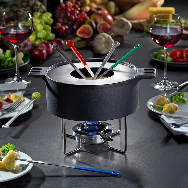 Veilig ondersteund door de fijne metalen standaard, lijkt de massieve gietijzeren pot boven de tafel te zweven. Mono design fonduepan op tafel, omringd door ingrediënten en wijn.