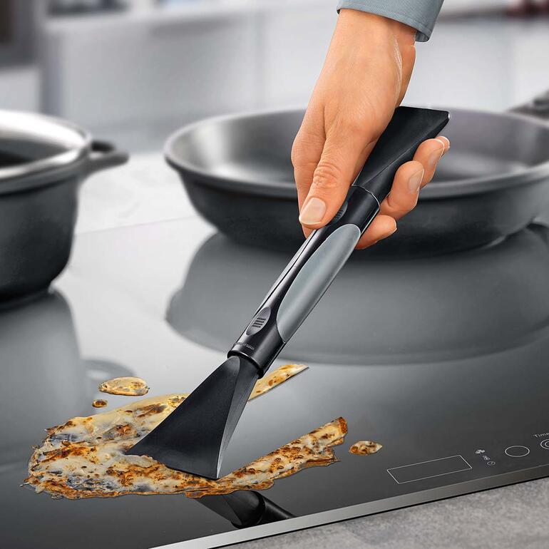 Hand die krastvrije kookplaat schraper 2-in-1 op een zwart kookoppervlak houdt.