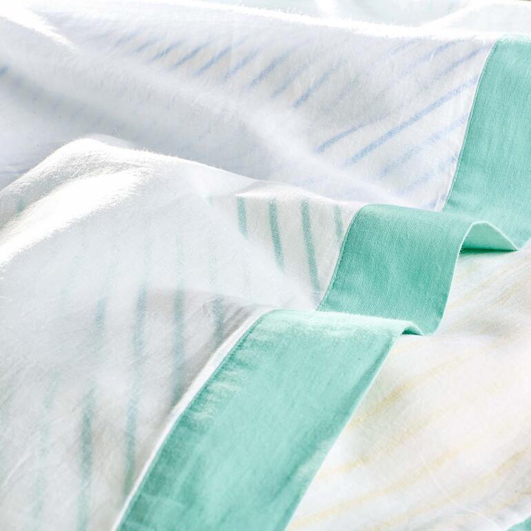 Gemaakt met drie lagen: boven- en onderkant zijn gemaakt van iets transparant katoenweefsel; de middelste laag in een kleurrijk streepdessin schijnt doeltreffend door. Close-up van de Breezyblanket met een groene rand.
