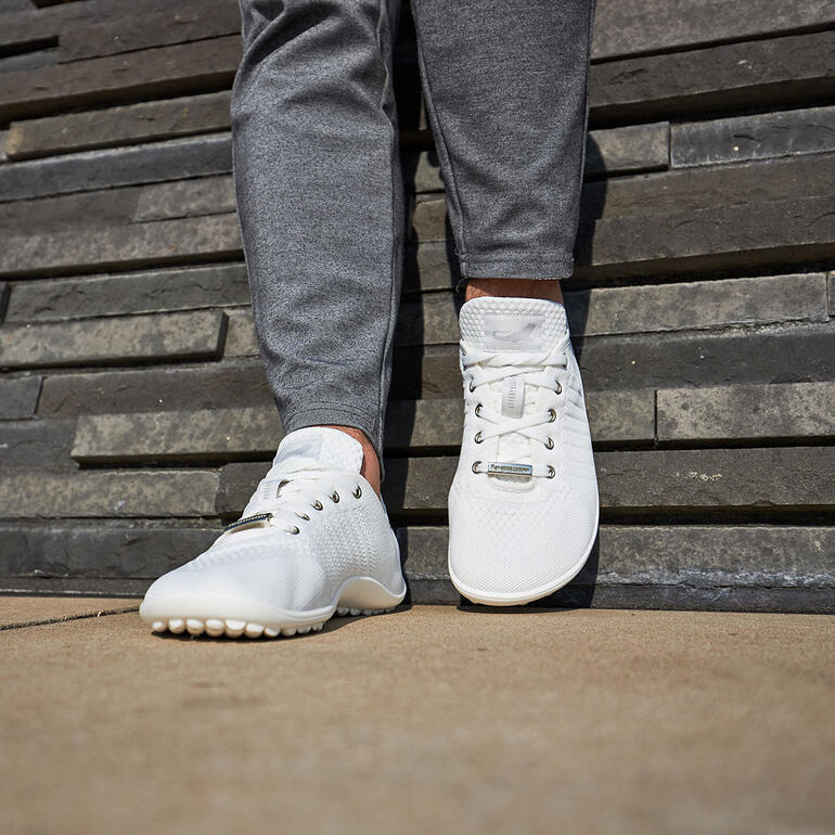 Persoon in grijze joggingbroek staat voor een stenen muur met witte sneakers.