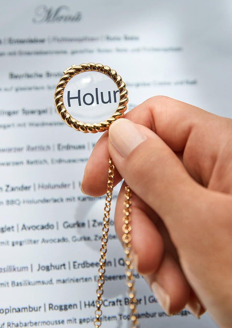 Hand houdt vergrootglas aan een gouden ketting boven een menu.
