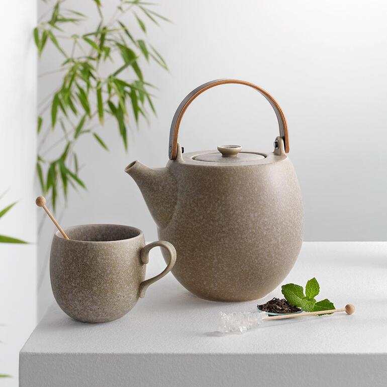Theepot en beker in grijs keramiek op een tafel met een plant op de achtergrond