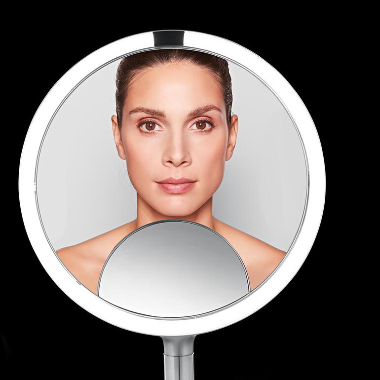 Bekijk het algemene effect van uw make-up in de 1:1-weergave, gebruik de spiegelzijde met 5x vergroting bijvoorbeeld om eyeliner en lipliner te corrigeren en gebruik het venster met 10x vergroting om de kleinste details te controleren. Close-up van een cosmeticaspiegel met het gezicht van een vrouw