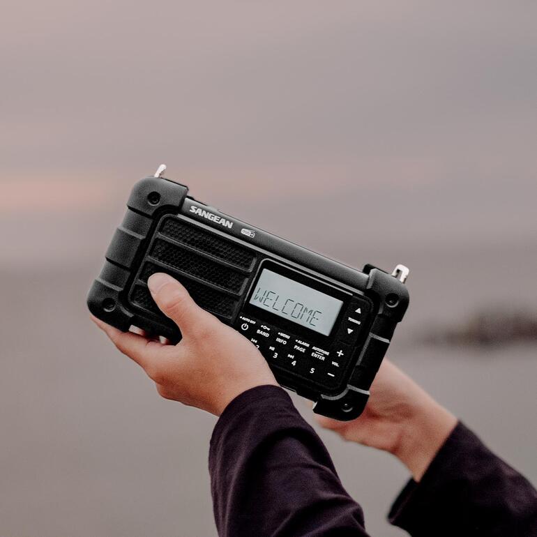 Persoon houdt digitale survivalradio omhoog terwijl hij/zij op het strand staat.