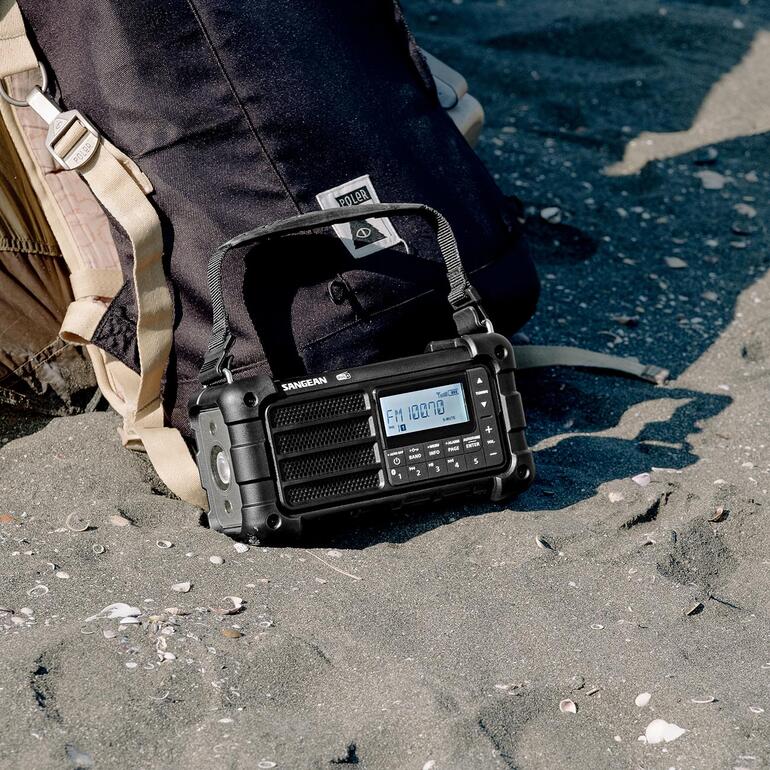 Digitale survivalradio staat in het zand naast een rugzak met een drager op de rug.