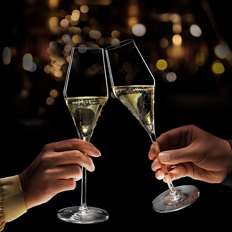 Wanneer de glazen worden opgetild, schijnt een gloed van licht op het fonkelende spel van bubbels in de glazen. Twee verlichte champagneglazen klinken