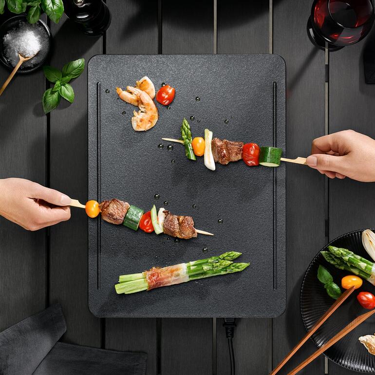 Dankzij het innovatieve, 5-laagse verwarmingssysteem wordt het grilloppervlak optimaal gelijkmatig verwarmd. Handen steken spiesjes met groenten en vlees op de tafelgrill.